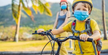 Active school kid boy and his mom in medical mask and safety helmet riding a bike with backpack on sunny day. Happy child biking on way to school. You need to go to school in a mask because of the coronavirus epidemic.