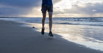 Exercising outside. A jogger in lightweight blue long sleeve t-shirt, shorts and running sneakers trains to marathon, running across the Baltic Sea beach, on cloudy windy rainy early morning at down